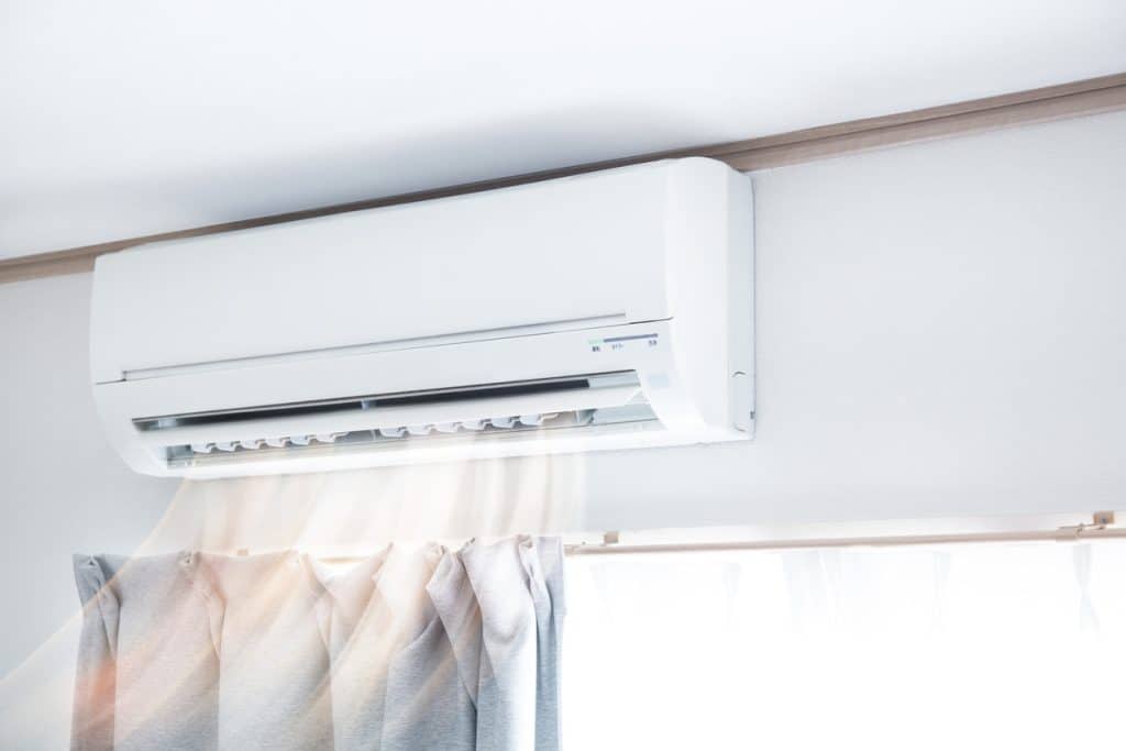 Wall-mounted split air conditioner blowing cool air into a bright room above a window with curtains.